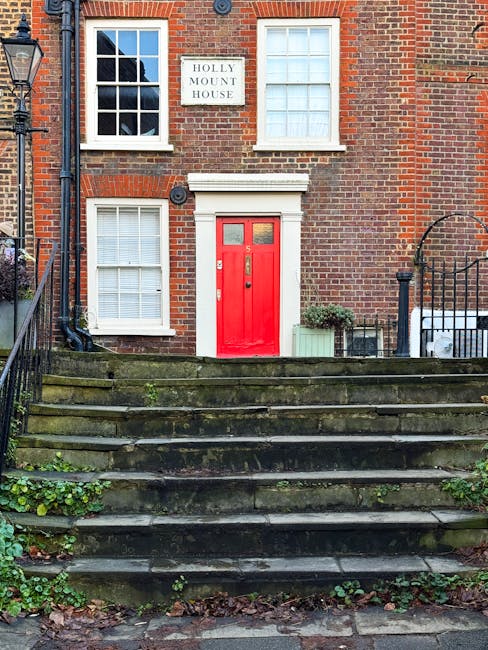 The front exterior of a brick terraced house with a bright red door set into a white frame, situated at the top of a flight of weathered stone steps covered in moss and fallen leaves. To the left of t