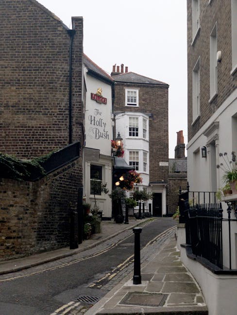 A residential street in West Hampstead with parked cars lining both sides of the narrow tarmac road. The vehicles include small vans, larger delivery trucks, and passenger cars, some with protective c
